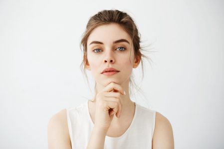 Portrait of young beautiful brunette girl looking at camera posing touching face over white background. Copy space.